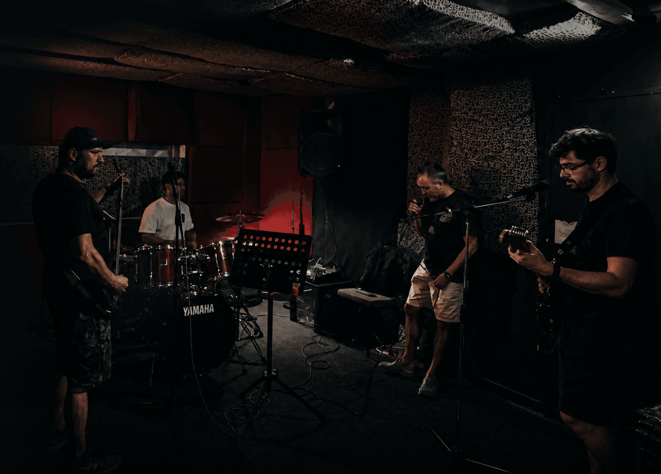 Four piece band rehearsing in a low light studio: bassist in a cap at left, drummer at the red Yamaha kit behind, vocalist at a stand mic in the centre, second guitarist with glasses at right. Red acoustic panels, patterned pillar, foam treated ceiling.